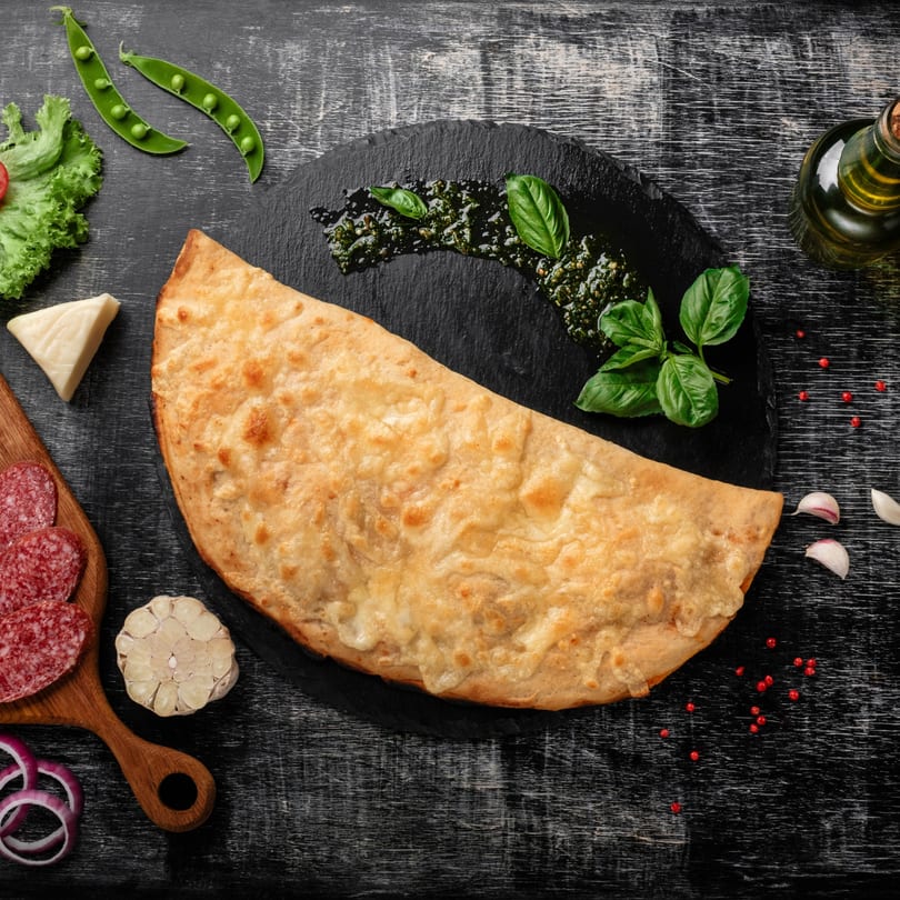 Wacky Zaki's calzone with pesto, basil, salami, cheese, and vegetables on a dark wooden table.