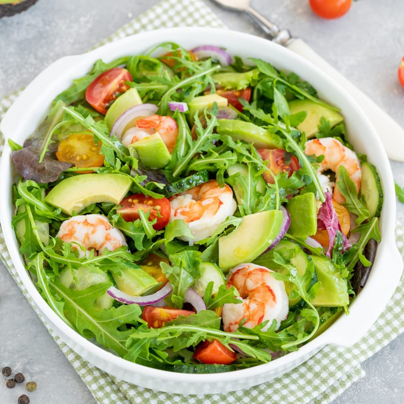 A vibrant shrimp salad with avocado, cherry tomatoes, red onion, and arugula in a white bowl.
